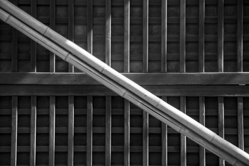 Black and white image of a wooden ceiling with a grid pattern. Two long beams run diagonally across the frame, intersecting the grid lines and casting shadows. The texture and lines create an abstract geometric design.