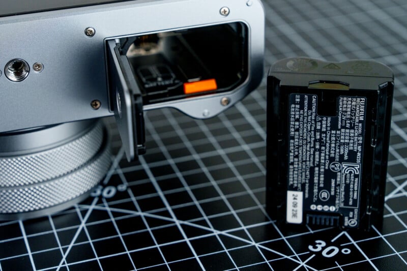 A camera with an open battery compartment next to a removed battery. The camera is placed on a black grid-patterned surface with a visible "30°" marking.