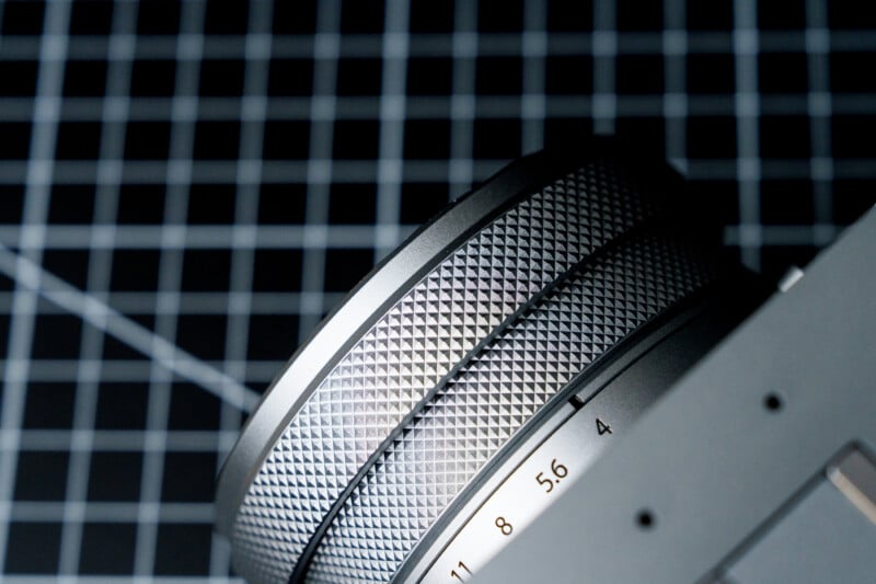 Close-up of a camera lens with a textured metal focus ring. The background features a black grid pattern.