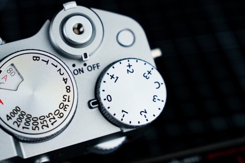 Close-up view of a silver camera's top dials. The left dial shows shutter speed settings and a meter, while the right dial features exposure compensation numbers. The surface has a textured grip, set against a black grid background.