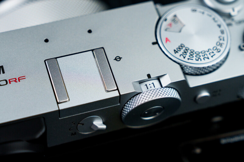 Close-up of a retro-styled silver camera top featuring a shutter speed dial, a textured adjustment knob, and a hot shoe mount. The design includes engraved numbers and letters, giving it a classic and mechanical aesthetic.