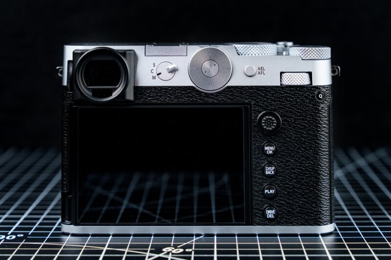 A vintage-style digital camera with a metal top and black leather grip is shown from the back. The screen and control buttons, including a menu and playback button, are visible. The camera is placed on a grid-patterned surface.