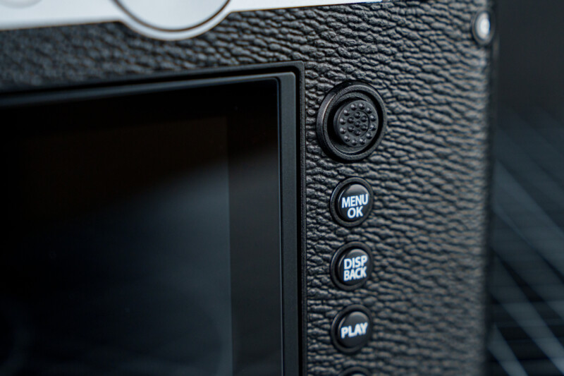 Close-up of a camera's backside showing a textured grip and several buttons: a circular button with a textured surface, and three labeled buttons: "Menu/OK," "Disp/Back," and "Play," next to an LCD screen.