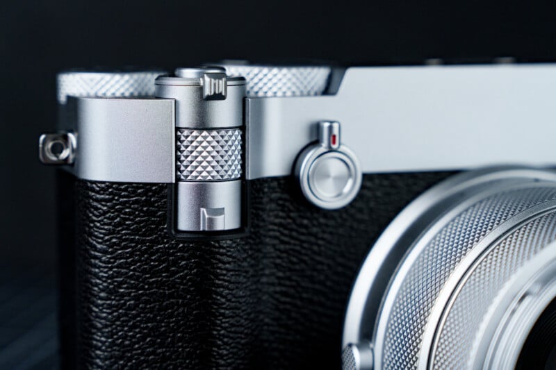 Close-up of a silver and black camera, focusing on its textured metal dials and lens. The camera features a sleek design with a small red dot button and a textured grip. The background is a dark gradient, emphasizing the camera's details.