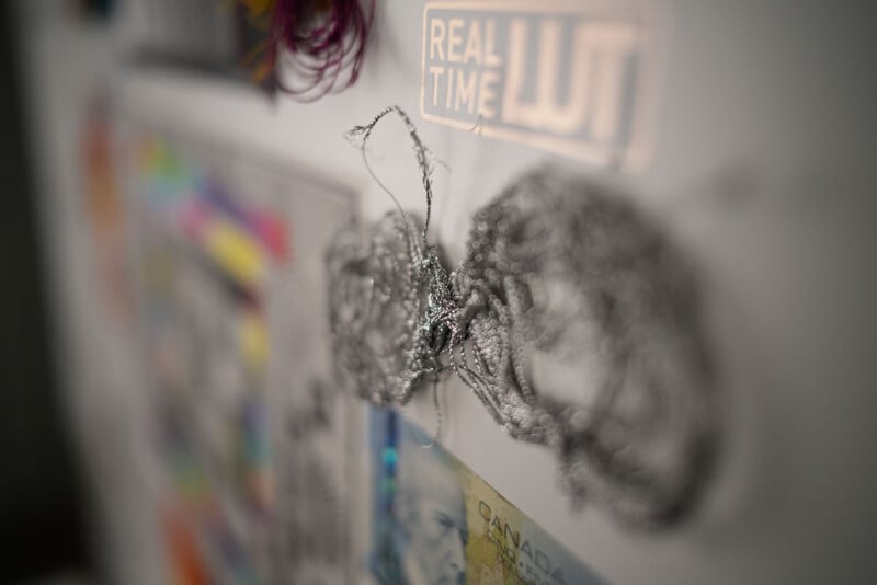 Close-up of a tangled wire sculpture affixed to a wall, with blurred colorful artwork and a sticker reading "REAL TIME LUT" in the background. The image is slightly out of focus.