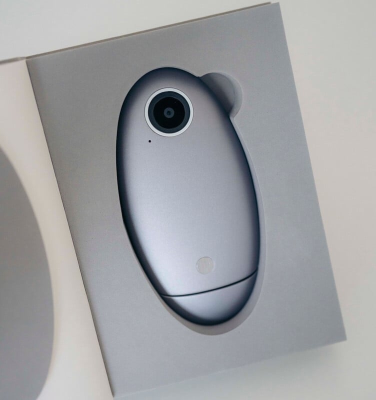 A sleek, silver, oval-shaped camera device with a single lens is placed inside a custom-cut gray box. The minimalist packaging and smooth design give the device a modern, high-tech appearance.