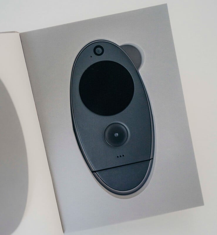 A photo of an open booklet showing an image of an oval-shaped electronic device with a camera, a circular touchpad, and a button, set against a plain background.