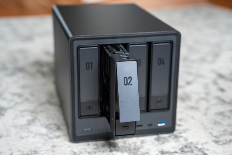 A black network attached storage (NAS) device with four drive bays sits on a speckled countertop. The second bay, labeled "02," is partially pulled out while the others remain closed.