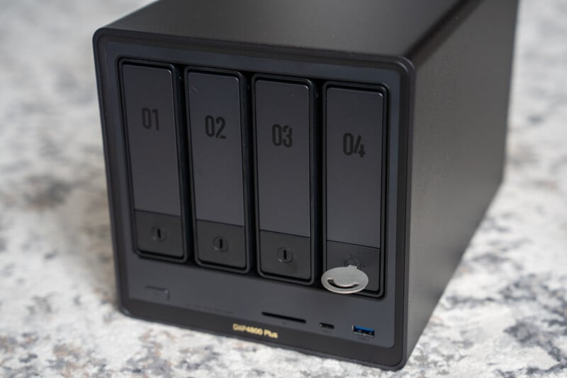 A black network-attached storage (NAS) device with four numbered drive bays, labeled 01 to 04, sits on a light-colored surface. One bay has a key inserted in its lock.