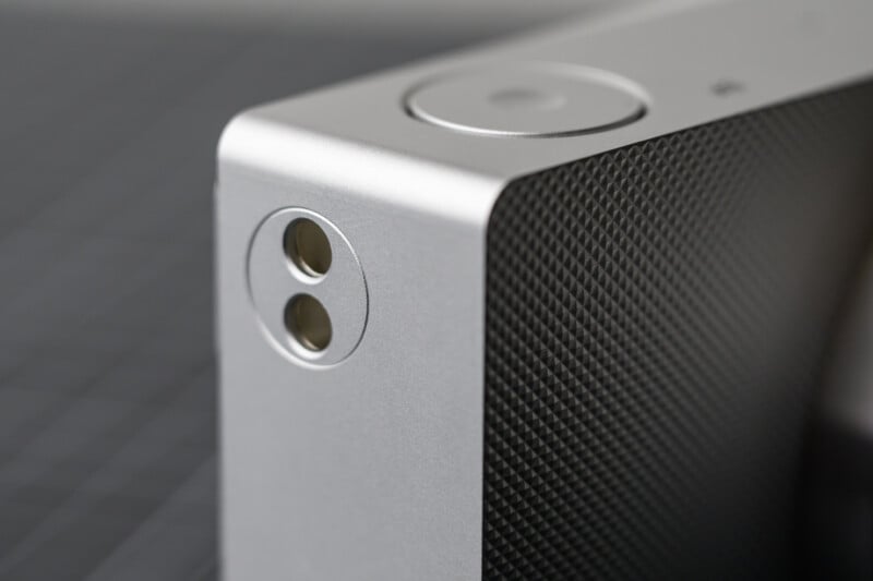 Close-up of a sleek, silver portable speaker corner. Features a circular button and dual input/output ports on a textured surface. The design is minimalist, with a matte finish and subtle detailing.