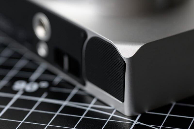 Close-up of a sleek electronic device, possibly a computer or gaming console, resting on a black grid-patterned surface. The device has a metallic finish and vents on the side.