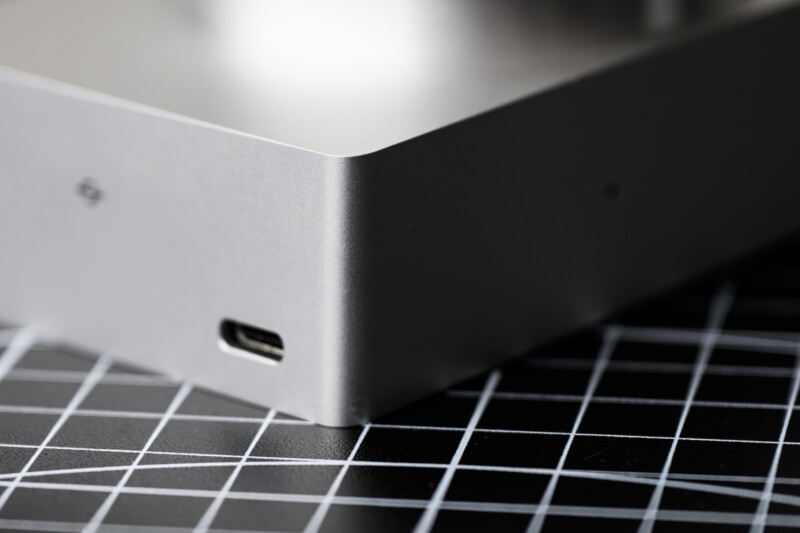 Close-up of a sleek, silver electronic device corner with a USB-C port. The surface is smooth and metallic. The device is resting on a black and white grid-patterned surface.