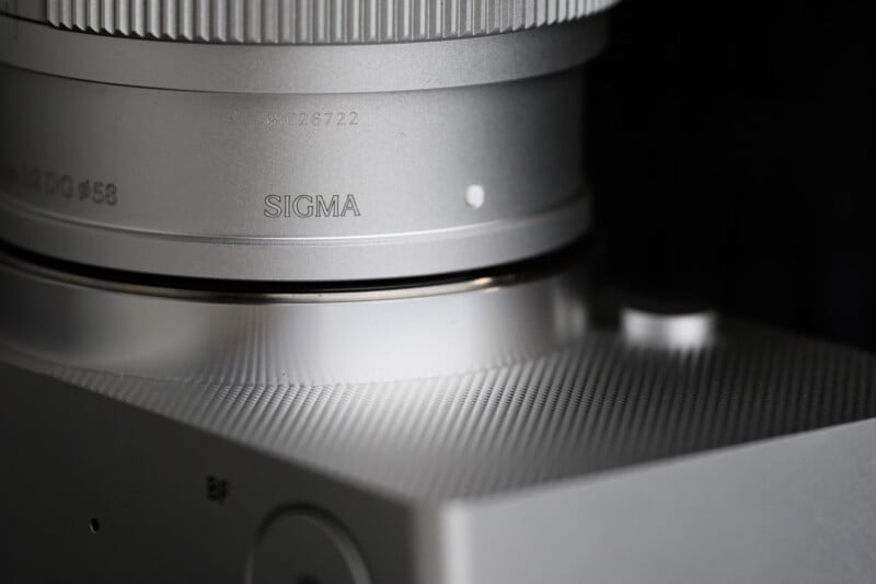 Close-up view of a silver Sigma camera with a textured surface. The image focuses on the lens and a portion of the camera body, highlighting the engraved Sigma logo and serial number.