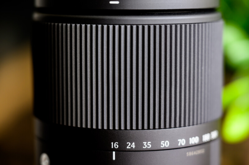 Close-up of a black camera lens with a focus on the textured zoom ring. White numbers indicating focal lengths ranging from 16 to 100 are visible. The background is blurred with a hint of green.