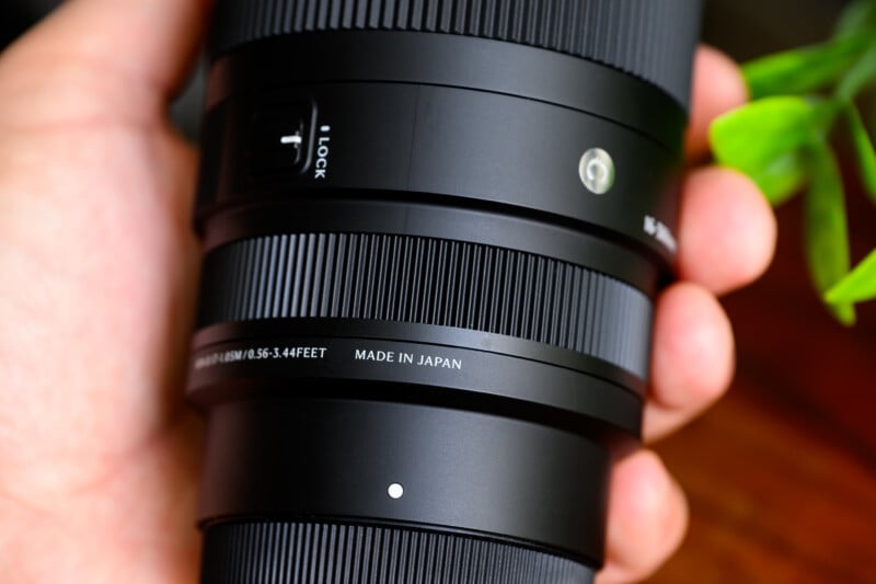 Close-up of a person's hand holding a black camera lens with visible text stating "Made in Japan" and specifications, featuring a lock switch and green plant leaves in the background.