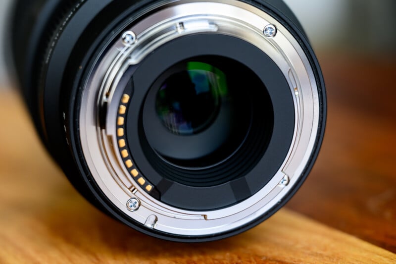 Close-up of a camera lens resting on a wooden surface, viewed from the rear mount side. The lens's electronic contacts and mount screws are visible, along with the glass elements. The focus ring and body are also partially visible in the background.