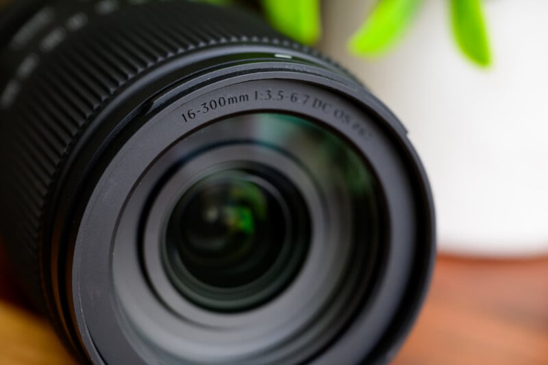 Close-up of a camera lens with "16-300mm 1:3.5-6.3 DC OS" labeling. The lens is black with fine ridges for grip, and a blurred green plant in the background adds a touch of color.