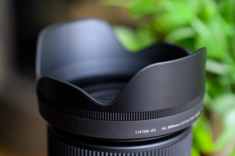Close-up of a black camera lens with a petal-shaped hood. The focus is on the lens's textured surface with engraved details. In the blurred background, there is a hint of greenery.