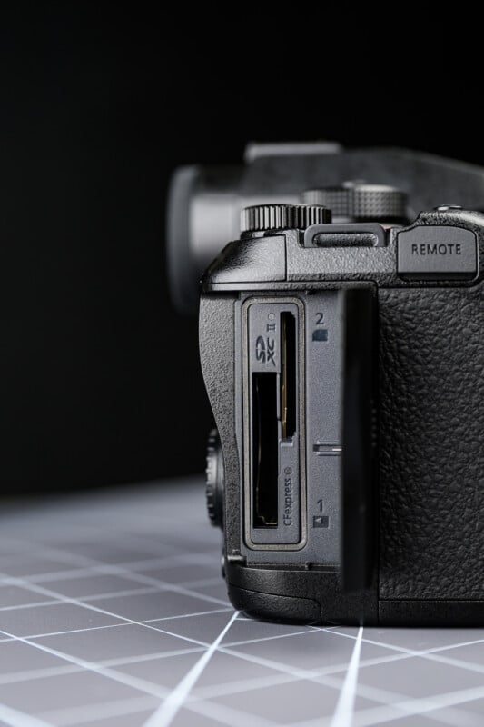 Close-up of a digital camera showing two SD card slots labeled 1 and 2, with one SD card partially inserted. The camera is placed on a checkered surface with a black background.