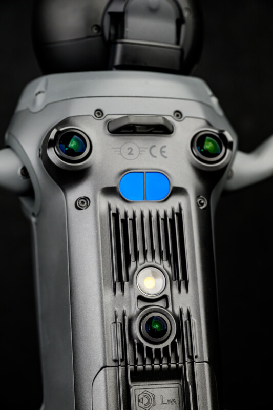 Close-up view of the front of a black and gray drone, showing its sensors, cameras, and a blue button against a dark background.