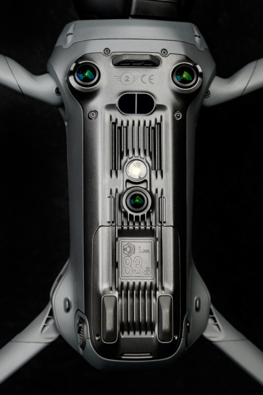 Close-up view of the underside of a gray drone, showing camera sensors, vents, and technical markings, set against a black background.