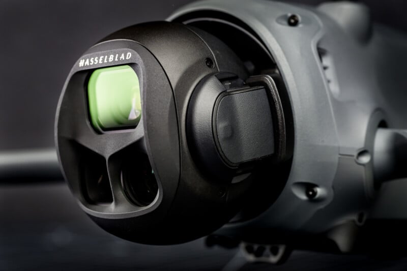 Close-up of a Hasselblad camera mounted on a grey drone, showing the lens and detailed camera housing. The background is dark and out of focus, highlighting the camera’s intricate design.