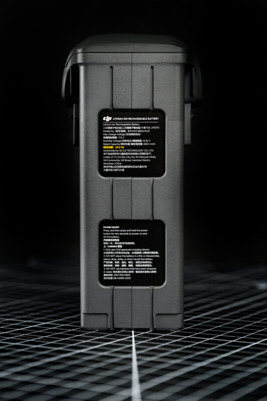 A close-up of a DJI lithium-ion rechargeable battery standing upright on a grid-patterned surface, with detailed warning and usage labels visible on the front.