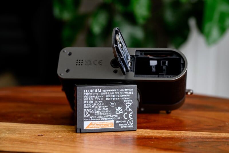 A Fujifilm rechargeable lithium-ion battery is placed in front of a camera with its battery compartment door open, resting on a wooden surface with a blurred green background.