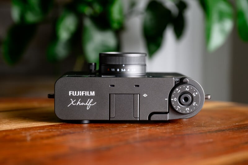 Top view of a black Fujifilm X-H2 camera resting on a wooden surface, showing the lens, control dials, and Fujifilm branding, with green leaves blurred in the background.