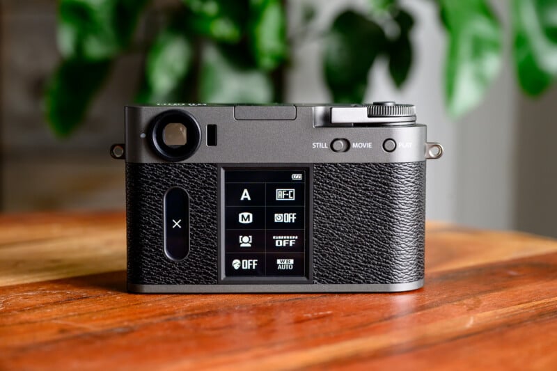 A digital camera with a textured grip sits on a wooden surface. Its display shows various shooting mode icons and settings. Green plants are blurred in the background.