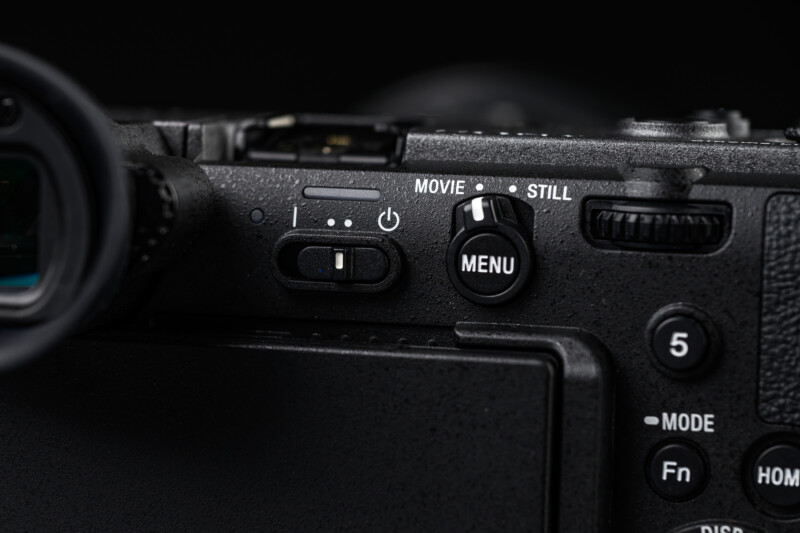 Close-up of a black digital camera showing the power switch, movie/still mode toggle, menu button, and part of the control dial against a dark background.