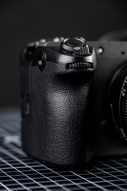 Close-up of a black camera body with a textured grip and control dials, placed on a grid-patterned surface with a blurred dark background.