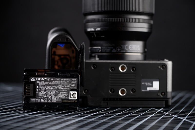 A close-up of a camera placed upside-down on a grid-patterned surface, with its battery compartment open and a Sony NP-FZ100 battery pack partially removed.