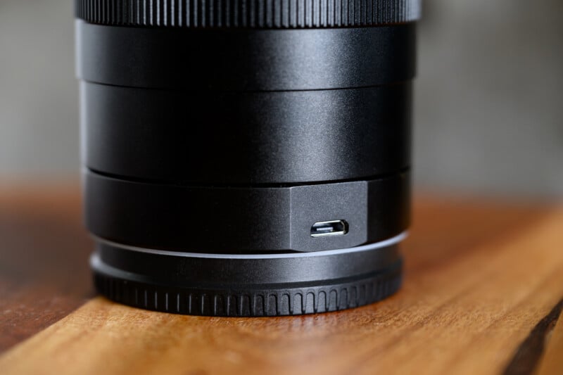 Close-up of a black camera lens placed on a wooden surface, with a visible micro-USB port on the side of the lens barrel.