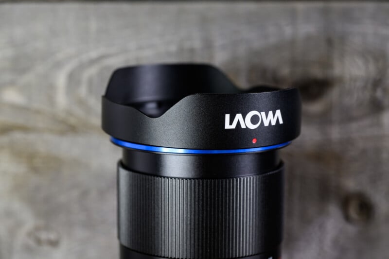 Close-up of a black Laowa camera lens with a blue ring and a lens hood, set against a blurred wooden background. The brand name "Laowa" is visible in white on the lens hood.