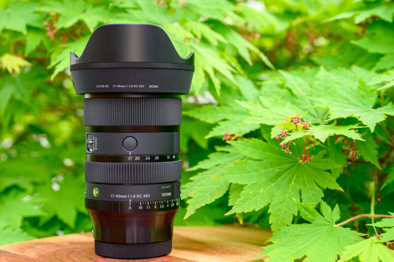 A Sigma 17-40mm camera lens stands upright on a wooden surface outdoors, surrounded by green leaves in the background.