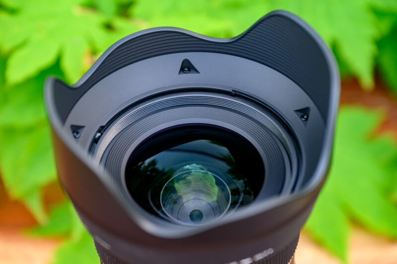 A close-up of a camera lens with a petal-shaped lens hood, showing clear glass elements and reflections, set against a blurred green leafy background.