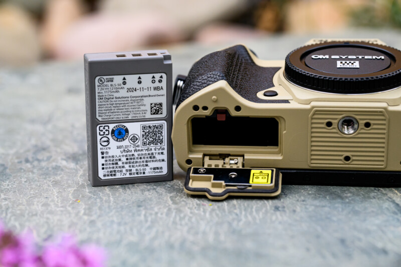 A close-up of a beige OM System camera with its battery compartment open, showing the battery slot. A camera battery stands upright beside the camera, displaying its label and details. The surface is stone-like with blurred flowers nearby.