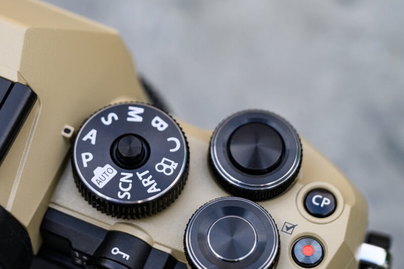 Close-up of a beige camera's mode dial and control knobs, showing various shooting modes like Auto, P, S, A, M, and custom settings, with textured black and silver dials.