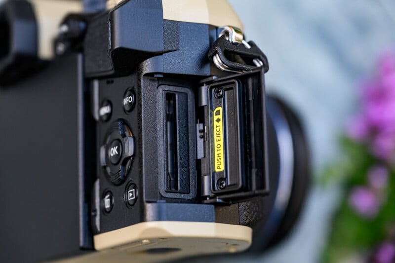 Close-up of a camera showing an open SD card slot with a yellow label reading "PUSH TO EJECT." Camera buttons and part of the lens are visible; background is blurred with a hint of pink flowers.