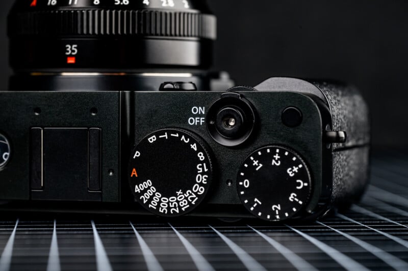 Close-up of a black digital camera showing the shutter speed dial, exposure compensation dial, and part of the lens with a 35mm focal length marking, set on a grid-patterned surface.
