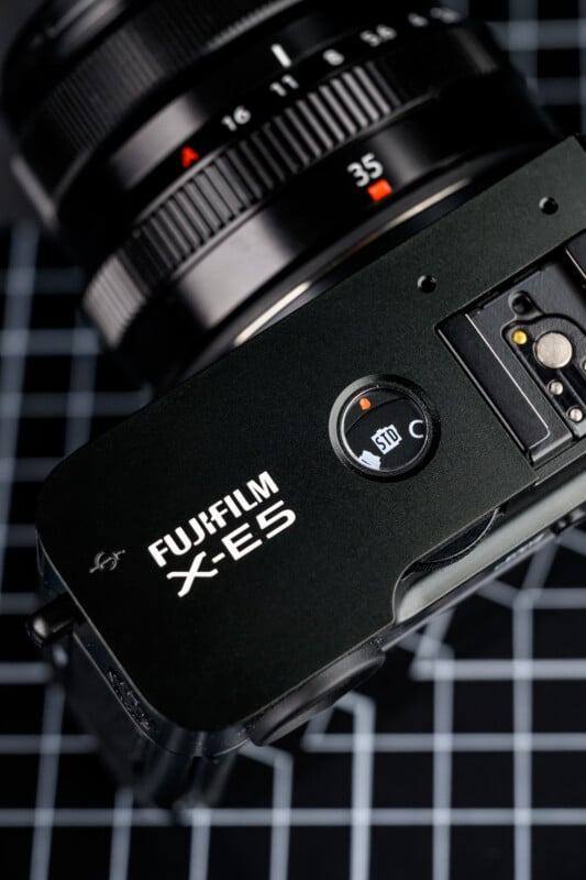 Close-up of a Fujifilm X-E5 camera placed on a grid-patterned surface, showing the top controls, lens with 35mm marking, and part of the camera’s branding.