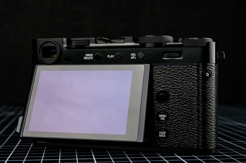 A close-up of the back of a black digital camera with a large LCD screen, several control buttons, and textured grip, set against a dark background with a grid-patterned surface.