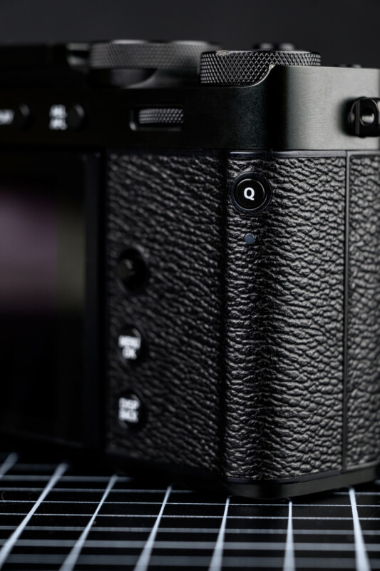 Close-up of a textured black camera body showing buttons and dials, with a "Q" button visible. The camera rests on a surface with a white grid pattern.