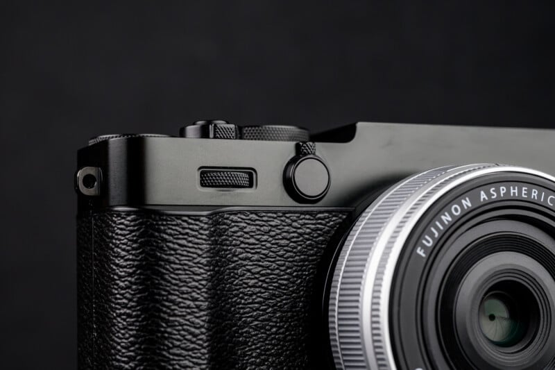 Close-up of a black Fujifilm camera, focusing on the textured grip, control buttons, and a portion of the lens with "Fujinon Aspherical" clearly visible against a dark background.