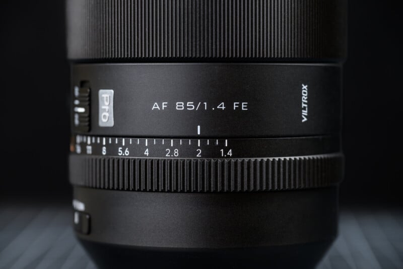 Close-up view of a Viltrox AF 85mm f/1.4 FE camera lens, showing the focus and aperture markings, textured focus ring, and product branding on a dark, blurred background.