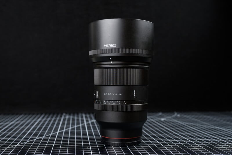 A Viltrox camera lens with a lens hood stands upright on a grid-patterned surface against a black background. The lens model "AF 85/1.4 FE" is visible on the side.