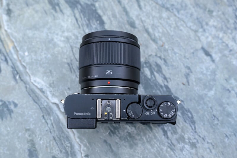 A top-down view of a Panasonic digital camera with a 25mm lens, placed on a grey marble surface. The camera's control dials and buttons are clearly visible.