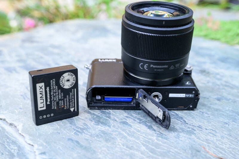 A Panasonic Lumix camera with its battery compartment open, showing the memory card inside. The camera battery is placed next to the camera on a stone surface, with greenery blurred in the background.