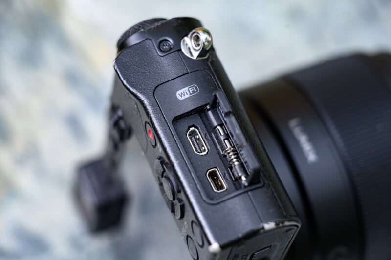 Close-up of a digital camera’s side, showing open port covers revealing HDMI, USB, and microphone input jacks, with a WiFi label above the ports. The camera lens is blurred in the background.